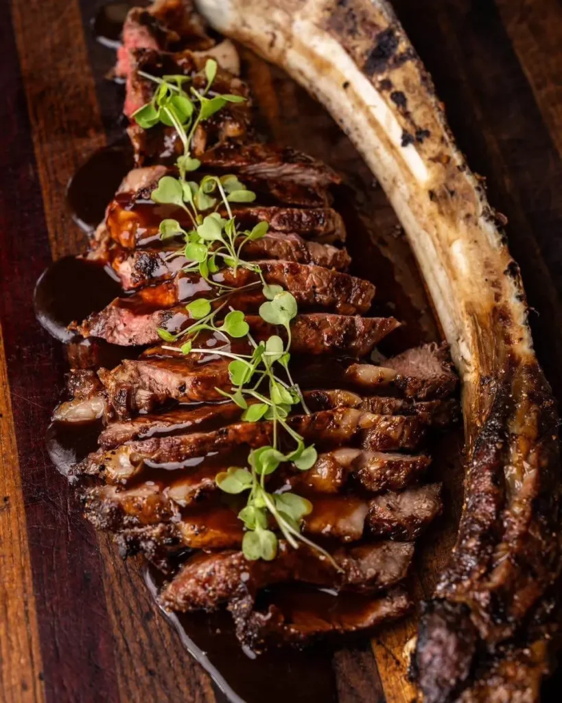 Sliced tomahawk ribeye fanned on a wooden board with fresh herbs at a catered dinner