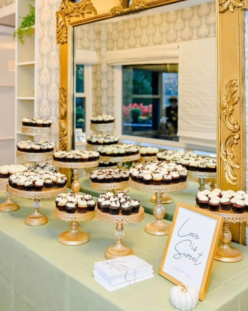 Tiered wedding dessert display with cupcakes on gold stands beside a gilded mirror at a catered reception